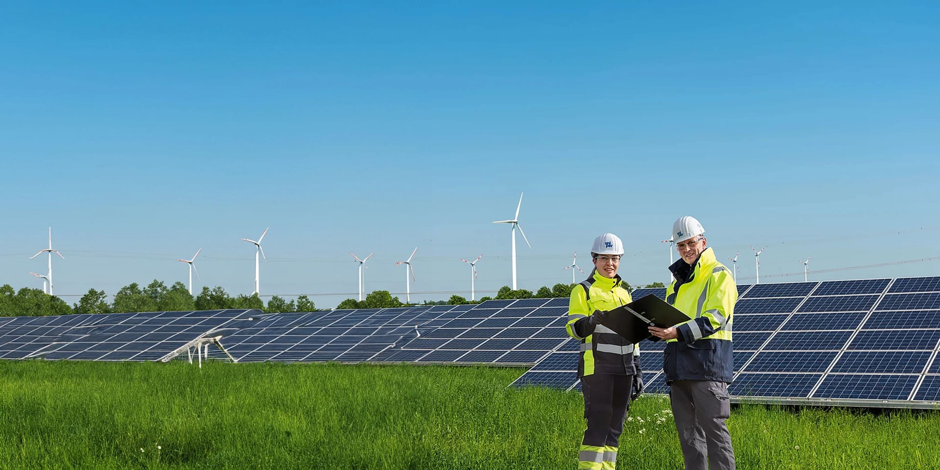 An employee stands in protective clothing in a field in front of a solar power plant, with wind turbines visible in the background.